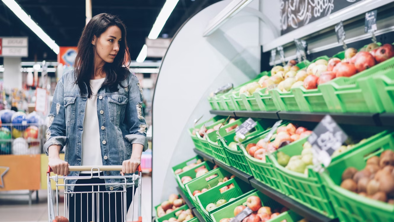 A woman pushing a cart and examining boxes of produce in a grocery store